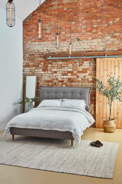 Industrial style bedroom with grey upholstered bed, white bedding, rustic brick wall, wooden sliding door, and modern pendant lighting