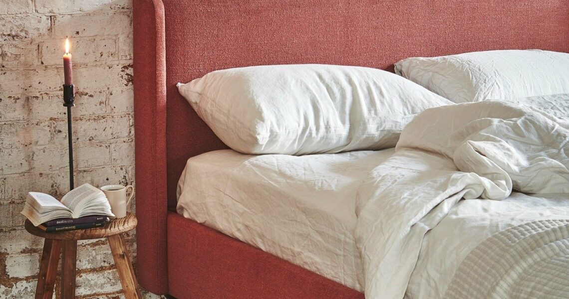 Rustic bedroom with red upholstered bed, white linen bedding, wooden stool nightstand, candle, and exposed brick wall